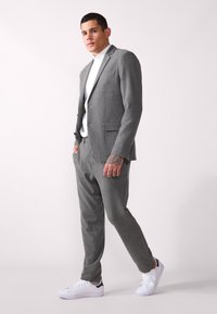 Grå skreddersydd dress med teksturert finish, featuring en tettsittende blazer og avsmalnende bukser, parret med en hvit skjorte og hvite sneakers.