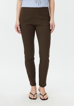Mujer con pantalones marrón oscuro ajustados, camiseta azul clara y sandalias tipo hilo negro, de pie frente a un fondo blanco.