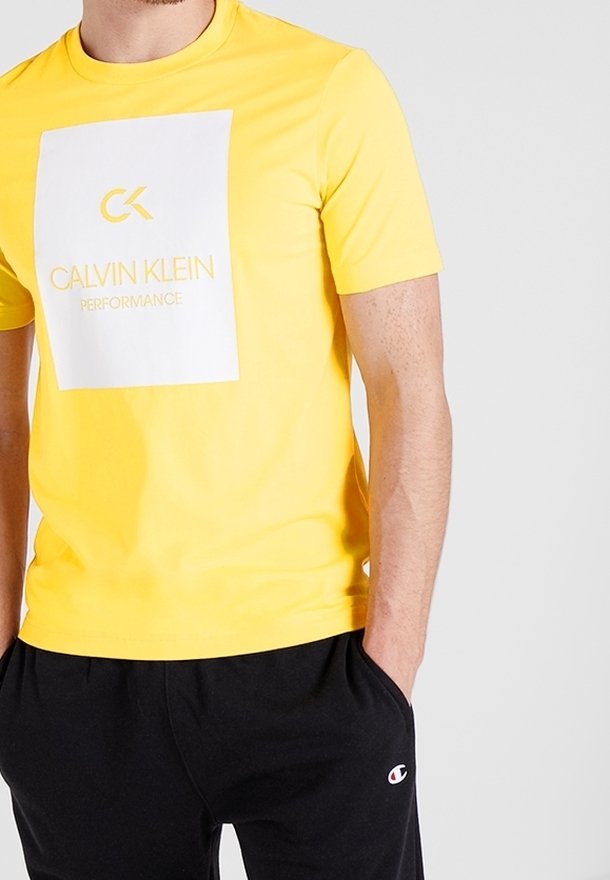 Homme portant un t-shirt jaune vif Calvin Klein Performance et un pantalon de survêtement noir Champion, les mains dans les poches, sur un fond blanc uni.