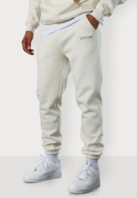 Weiße Baumwoll-Sweatpants mit elastischen Bündchen und seitlichen Taschen, mit einem kleinen schwarzen Logo. Kombiniert mit weißen Sneakers und sichtbaren weißen Socken.