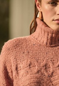 Ljusrosa stickad polotröja med texturerat mönster och ribbad krage. Materialet verkar mjukt med subtila inslag av blått.