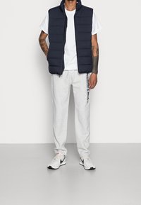 Gilet matelassé navy avec col haut, t-shirt blanc, pantalon de survêtement gris avec impression sur le côté, et baskets grises avec accents noirs.