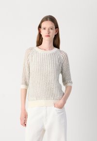 Witte gebreide trui met een losse structuur, 3/4 raglanmouwen, en geribbelde halslijn en zoom. Gecombineerd met witte broek, wat de textuur benadrukt.