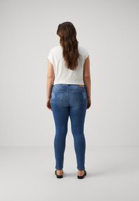 Blue fitted jeans with rear pockets, paired with a white short-sleeve top. The model is facing away, on a plain gray background.