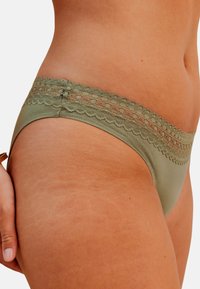 Culotte hipster en vert olive avec dentelle, présentant un corps en tissu lisse et des détails en dentelle festonnée le long de la taille.