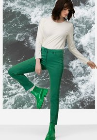 Zielone dżinsy w wąskim kroju z bocznymi szwami, zestawione z jas greenami gumowymi botkami do kostek i białą bluzką z długim rękawem.