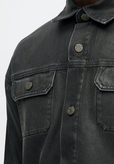 Temno siva denimska jakna z boutonskim sprednjim delom, dvema žepoma na prsih, kovinskimi gumbi in koničastim ovratnikom. Mehka tekstura in strukturirana zasnova.