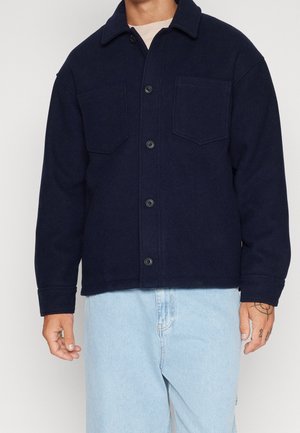 Mand iført navyblå knapjakke med to brystlommer over en beige skjorte og lyseblå jeans, der viser tatoveringer på venstre hånd.