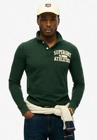 Dark green long-sleeve polo shirt with a collar and a white embroidered "Superdry Athletics" text. White knitted sweater tied at the waist.