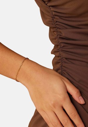 Close-up van een hand die rust op een geplooide bruine stof, met een delicate gouden schakelarmband om de pols.