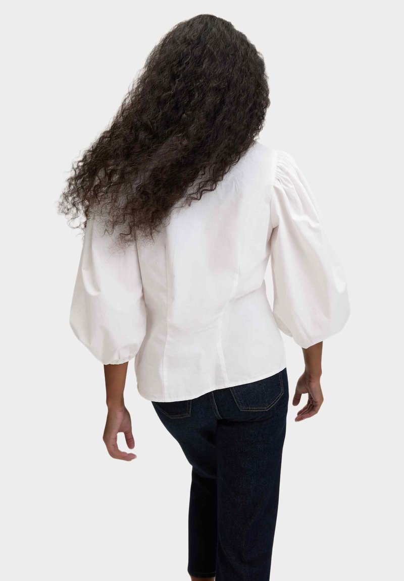 Witte blouse metpofmouwen en een aansluitende taille, ontworpen in een katoenmix. De achterkant heeft een gladde afwerking en een ronde zoom. Gecombineerd met donkere jeans.