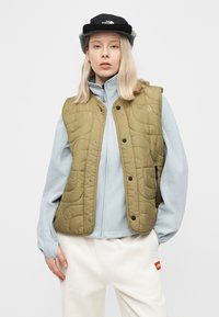 Γυναίκα που φοράει αμάνικο μπουφάν ελιάς πάνω από ανοιχτό μπλε φλις μπουφάν, λευκό παντελόνι και μαύρο καπέλο, στέκεται μπροστά σε απλό φόντο.