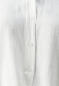 Camicia bianca con bottoni sul frontale, realizzata in un tessuto leggero e testurizzato con un sottile motivo intrecciato e bottoni in plastica trasparente.