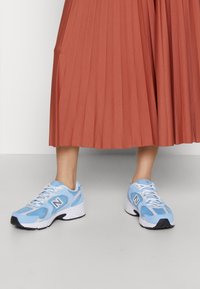 Jambes portant des baskets New Balance bleu clair et blanc avec des lacets blancs, associées à une jupe midi plissée orange rouille sur un sol clair.