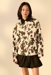 Hnědo-bílá bunda s cow printem, se dvěma předními kapsami, knoflíkovým zapínáním a černým límcem, nošena přes černou nadýchanou sukni.