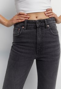 Jean noir taille haute en denim, avec une fermeture à bouton sur le devant, des accessoires en ton cuivré et deux poches avant.