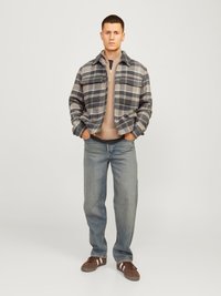 Plaid flanelinė striukė pilkos ir smėlio spalvų atspalviuose, su apykakle ir priekinėmis kišenėmis, dėvima ant smėlio spalvos megztinio ir šviesiai mėlynų džinsų, rudos spalvos batai.