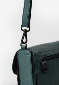 Borsa in pelle teal con una texture liscia, dotata di hardware nero, una tracolla removibile e una tasca con zip sul fronte.