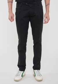Pantaloni neri in cotone con una vestibilità slim, design a cinque tasche, chiusura con bottone e zip, abbinati a sneaker bianche con dettagli verdi.