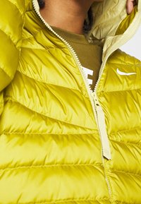 Kirkas limenvihreä untuvapusero, jossa on kevyt kangas ja tikattu kuosi. Varustettu täyskorkealla vetoketjulla ja valkoisella logolla.