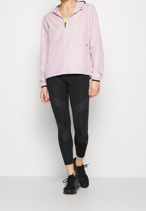 Ljust rosa zip-up jacka med huva, i kombination med svarta leggings som har geometriska prickmönster, och svarta träningsskor.