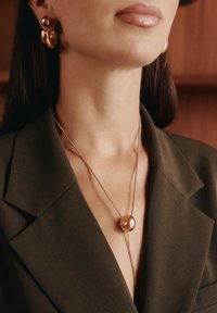 Collana in metallo color oro con un pendente rotondo, indossata sopra un blazer oliva con scollo a V profondo. Orecchini in oro abbinati con elementi circolari.