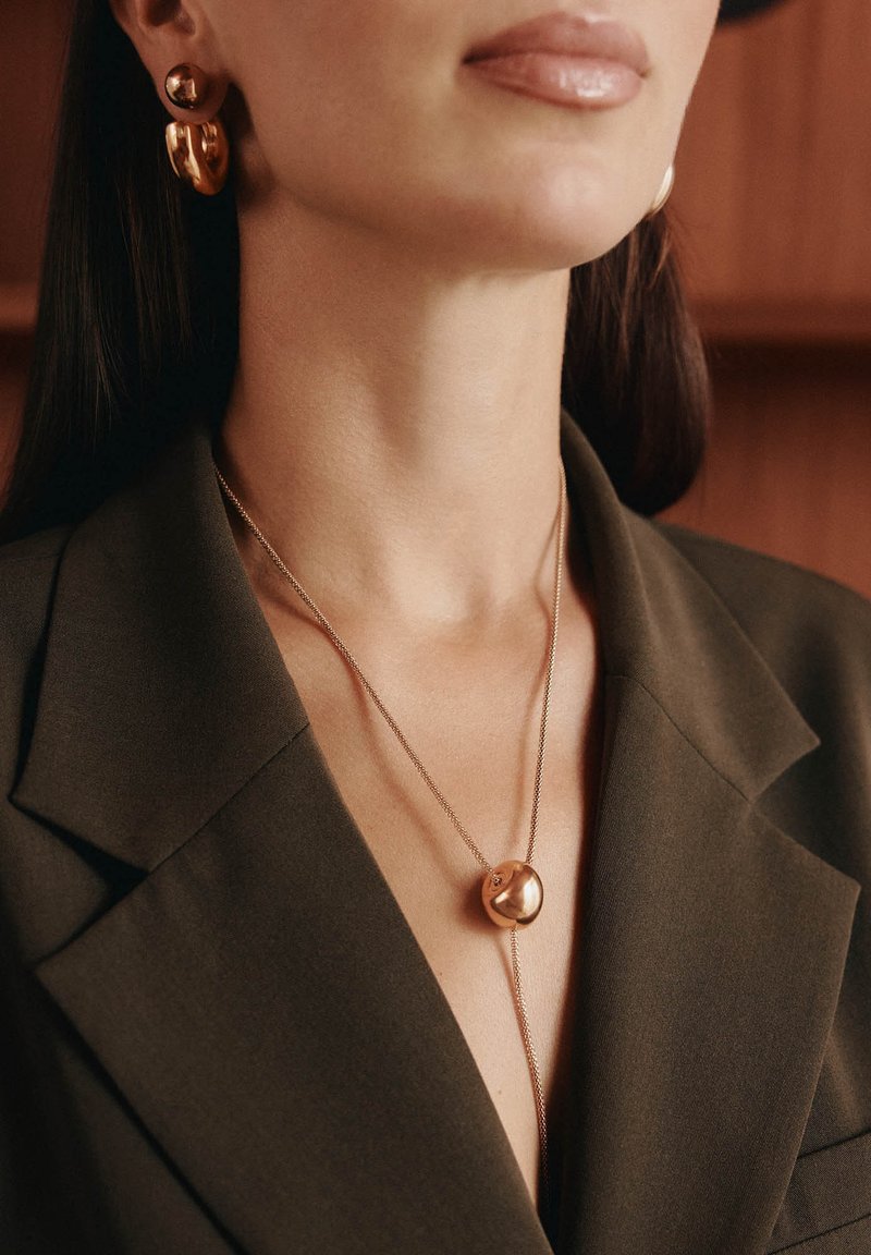 Collana in metallo color oro con un pendente rotondo, indossata sopra un blazer oliva con scollo a V profondo. Orecchini in oro abbinati con elementi circolari.
