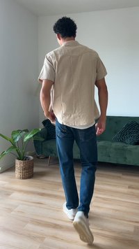 Rövid ujjú, bézs színű póló, lazább fazonnal, kék farmernadrág és fehér sportcipő, egy zöld kanapéval és cserépben lévő növénnyel berendezett szobában áll.