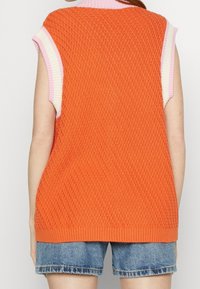 Henkilö, joka käyttää oranssia teksturoitua hihattomaa neuleliiviä vaaleanpunaisen ja valkoisen topin päällä sekä sinisiä farkkushortseja, kuvattu takaa.