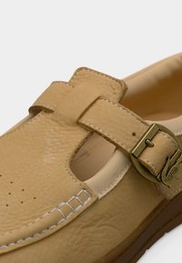 Chaussure en cuir fauve avec une bride à boucle dorée et des coutures blanches visibles sur un fond blanc.