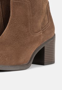Botine din suede maro, cu toc bloc, având accente cusute și panouri laterale pentru un design structurat. Talpa texturată pentru aderență.