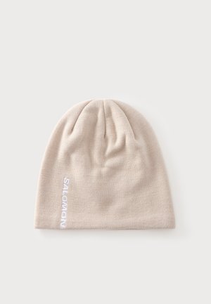 Bonnet en maille beige clair avec broderie verticale blanche du logo "SALOMON" sur le côté, présenté sur un fond clair uni.