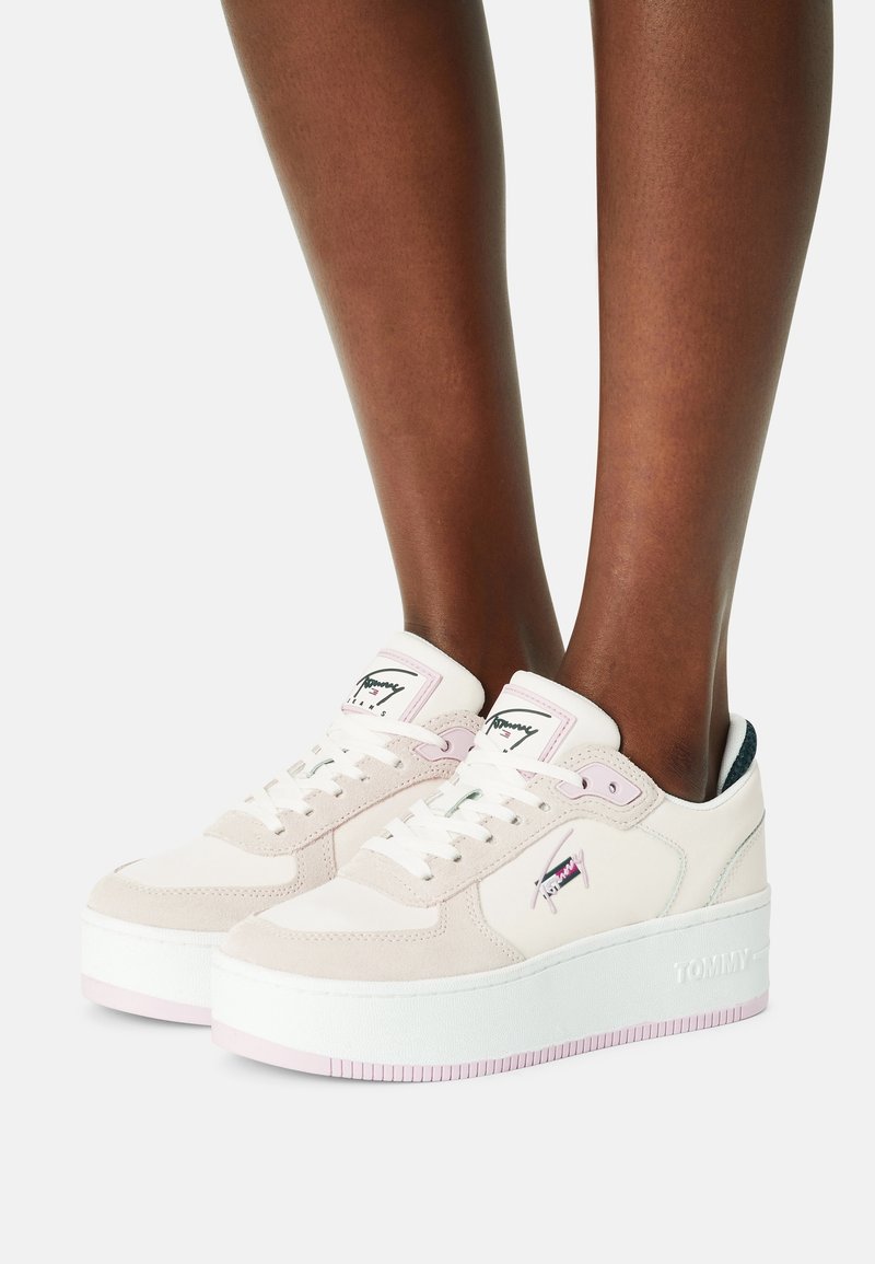 Baskets à plateforme blanches et rose clair avec lacets et logo, portées sur des jambes à la peau foncée, sur un fond clair uni.