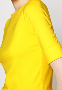 Gul bomullstop med rund halsringning och trekvartslånga ärmar med rullade manschetter, slät textur och figurnära siluett.