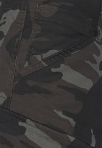 Tela con patrón de camuflaje en negro y gris, con costuras precisas y textura suave. Corte con bolsillos angulados y silueta ajustada.