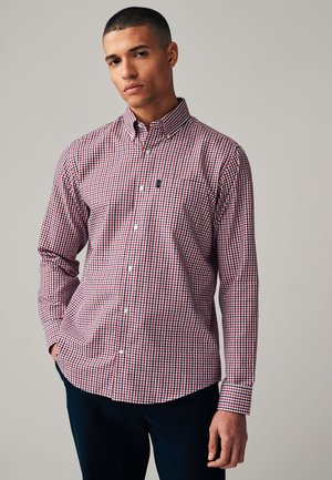 Homme portant une chemise à carreaux rouge, blanche et bleue à boutons, manches longues, et un pantalon foncé, debout devant un fond uni.