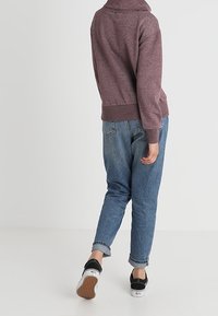 Persona con una sudadera color burdeos, jeans azules de puño y zapatillas negras y blancas caminando de espaldas sobre un fondo claro.