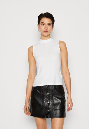 White sleeveless top with a high neckline and small red logo, paired with a black faux leather mini skirt featuring front buttons.