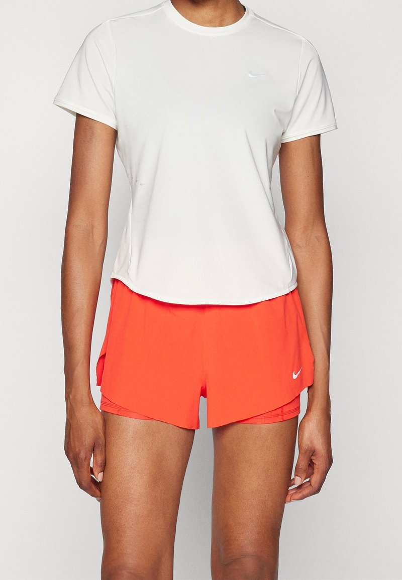 Camiseta deportiva blanca de manga corta combinada con pantalones cortos de color naranja brillante. Los pantalones cortos presentan un diseño en capas con un detalle del logo en el costado.