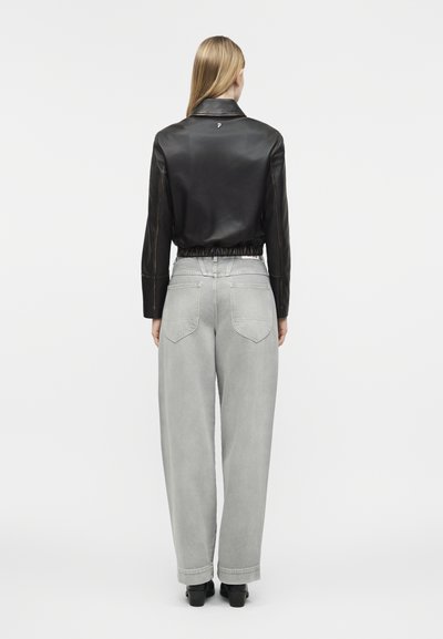 Woman with long blonde hair wearing a black cropped leather jacket and light gray wide-leg jeans, standing facing away on a white background.