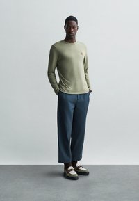 Olivno zeleni majica s dolgimi rokavi in majhnim logotipom, v kombinaciji s širokimi modrimi hlačami in večbarvnimi supergami. Minimalističen dizajn.