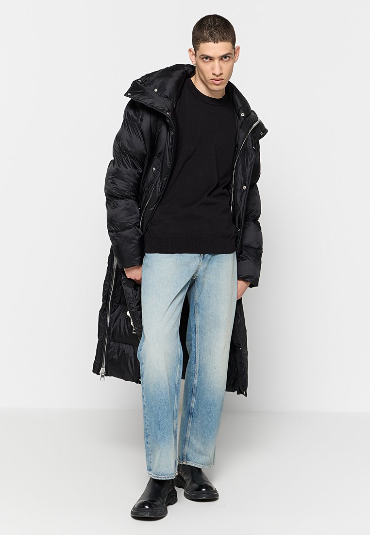 Schwarze gefütterte Jacke mit Kapuze, mit Reißverschluss vorne und seitlichen Taschen, kombiniert mit einem schwarzen Pullover und hellblauen geraden Jeans. Schwarze Stiefel.