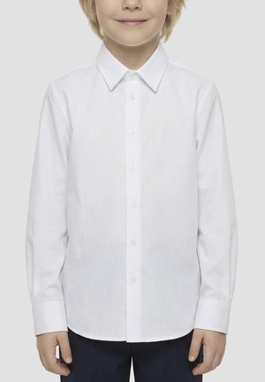 Kind draagt een netjes dichtgeknoopte witte overhemd met lange mouwen en een donkere broek, glimlacht lichtjes tegen een eenvoudige lichte achtergrond.