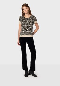 Top de manga corta con un intrincado diseño de paisley negro sobre un fondo crema, combinado con pantalones de terciopelo negro y botines de tacón cuadrado.