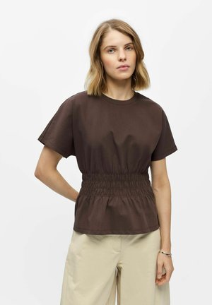 Vrouw met schouderlang blond haar, die een donkerbruine top met korte mouwen en geplooide taille draagt, en lichtbeige broek tegen een witte achtergrond.