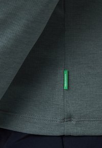 Gros plan sur un tissu texturé vert foncé avec une petite étiquette verte portant l'inscription « GREEN SHAPE » près d'une couture sur un vêtement foncé.