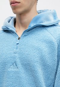 Giovane che indossa un pile zip-up di colore azzurro chiaro con un logo Adidas in rilievo sul petto e un colletto ampio.