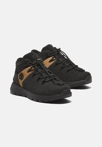 Chaussures de randonnée noires avec une tige en daim et synthétique, comprenant un panneau marron, des lacets en fil et une semelle extérieure en caoutchouc texturé.