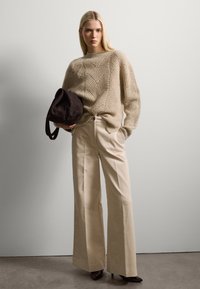 Beige gestrickter Pullover mit einem Zickzackmuster; cremefarbene, weit geschnittene Hose; dunkelbraune Wildleder-Schultertasche; spitze schwarze Absätze.