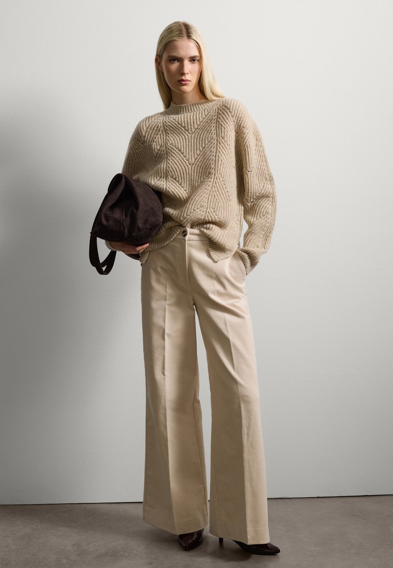Beige gestrickter Pullover mit einem Zickzackmuster; cremefarbene, weit geschnittene Hose; dunkelbraune Wildleder-Schultertasche; spitze schwarze Absätze.